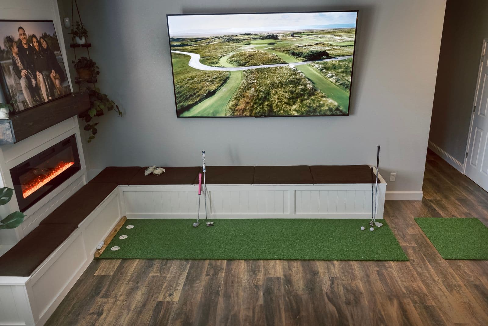 A home golf practice corner with a putting mat and a golf course displayed on a wall-mounted TV